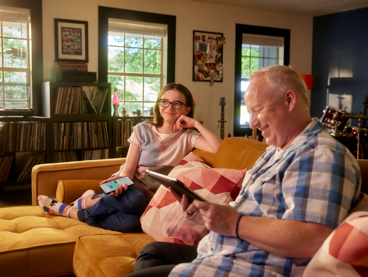 A girl and her dad sit on the couch while her dad smiles at a tablet. 