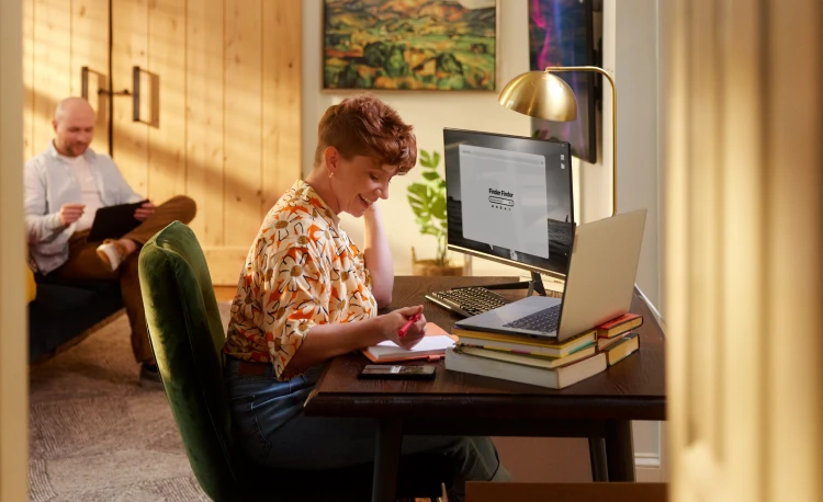 A woman sitting at her computer at her desk holds a pen over her notebook.