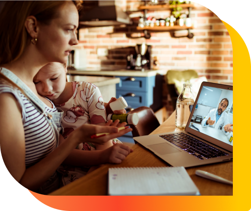 A woman sitting at a desk with a laptop open, video calling a medical professional, and holding a baby in her arms.